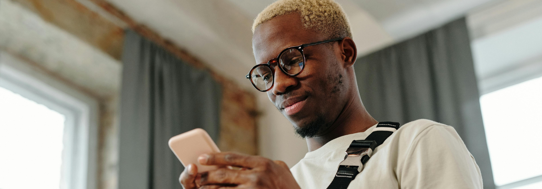 A black man with light hair and glasses looks at a phone in their hands. They appear to be smiling indoors.