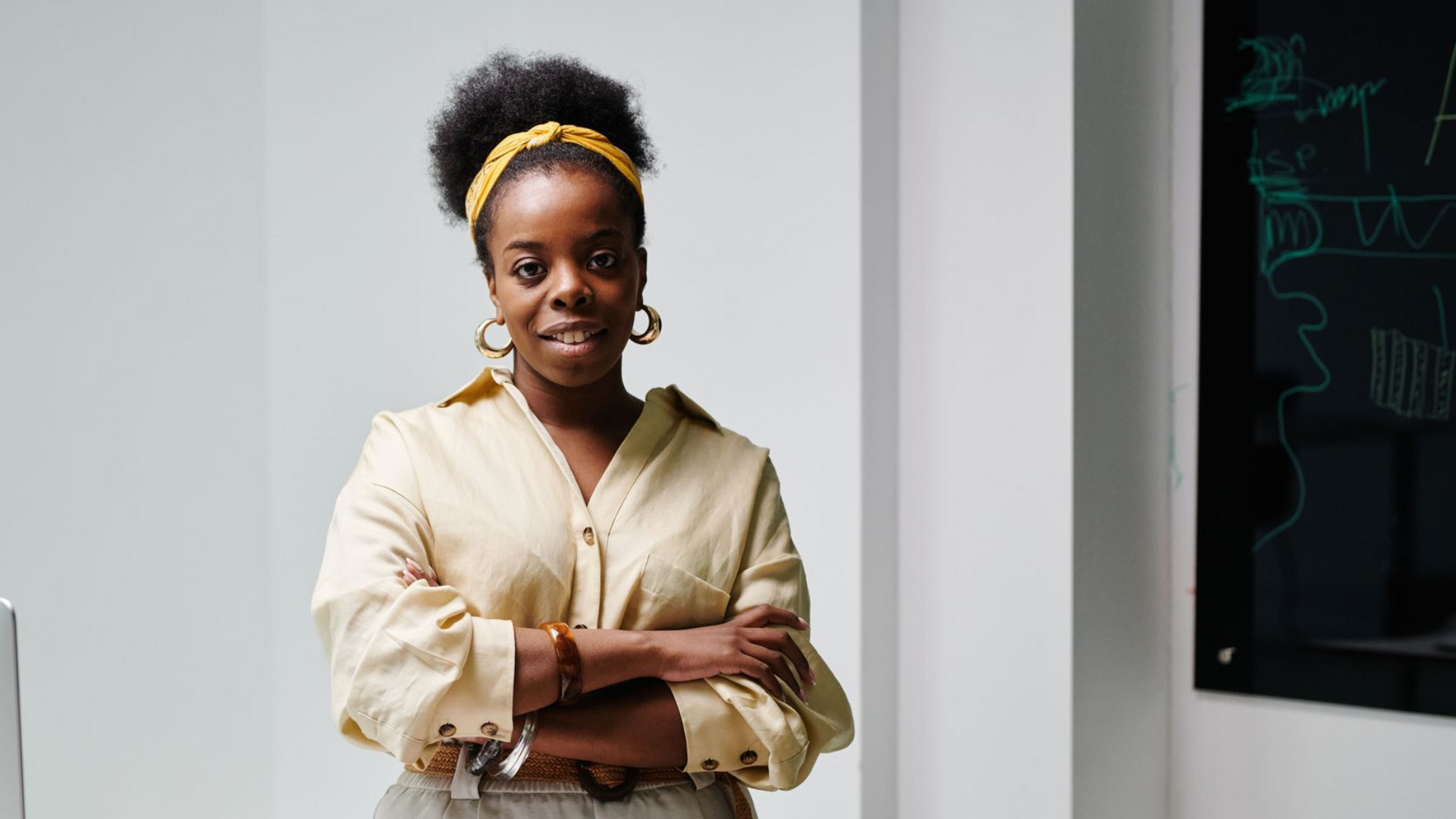 Person standing with arms crossed in a light blouse beside a white wall and chalkboard.