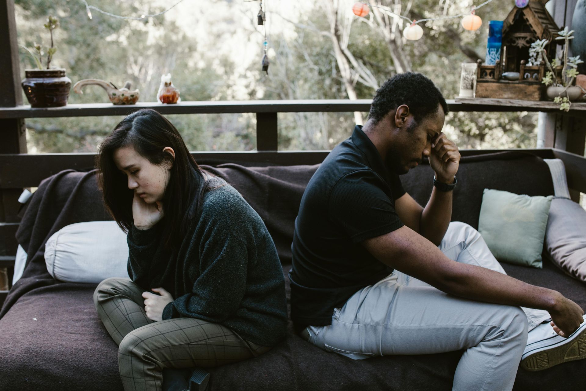 A couple sits apart on a couch, appearing distressed, outdoors on a porch.
