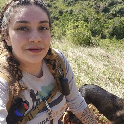 Leilah taking a selfie on a sunny trail with a dog and grassy hills in the background