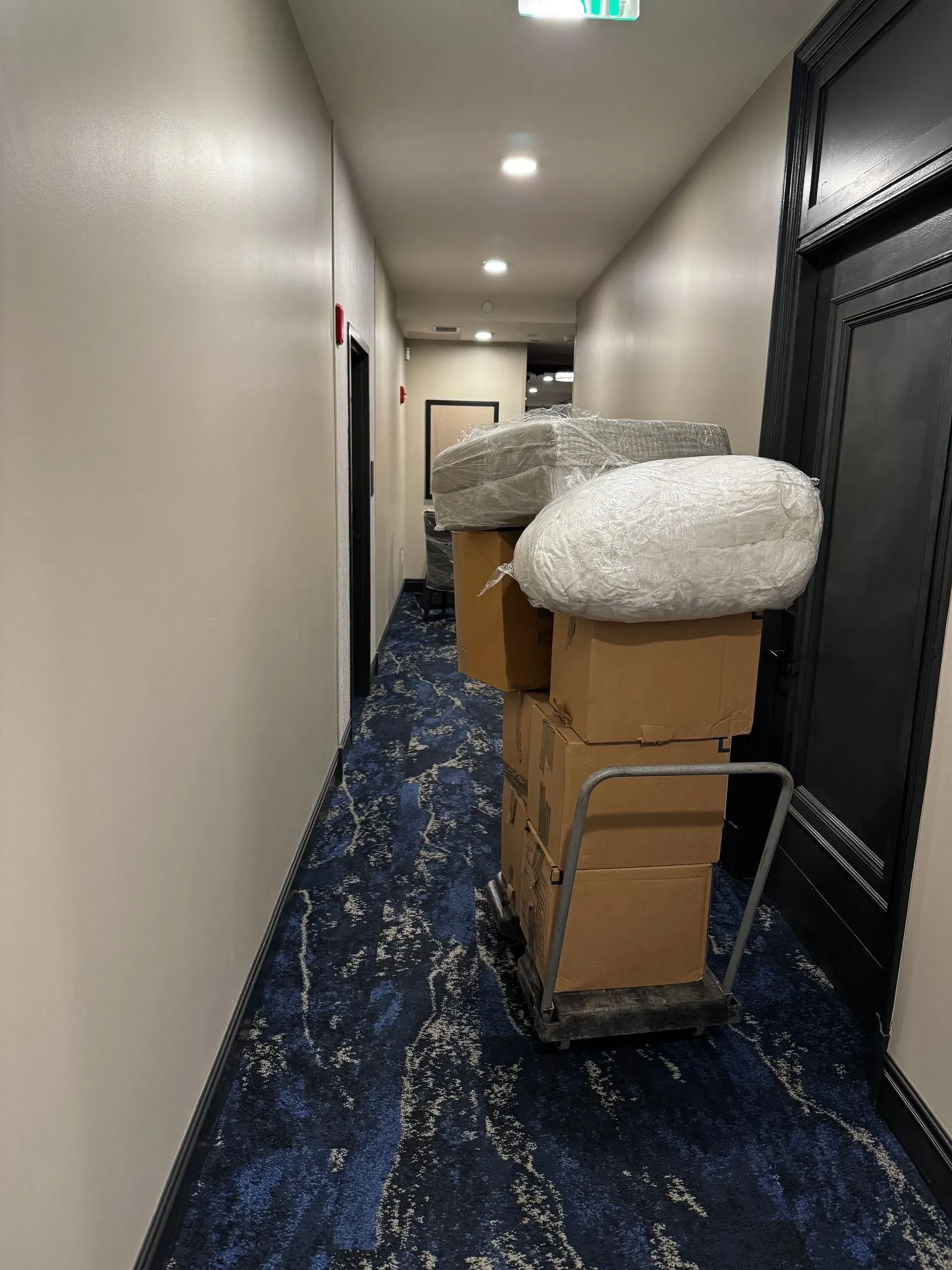 A hand truck loaded with boxes and bubble wrap in a hotel hallway with blue carpet.