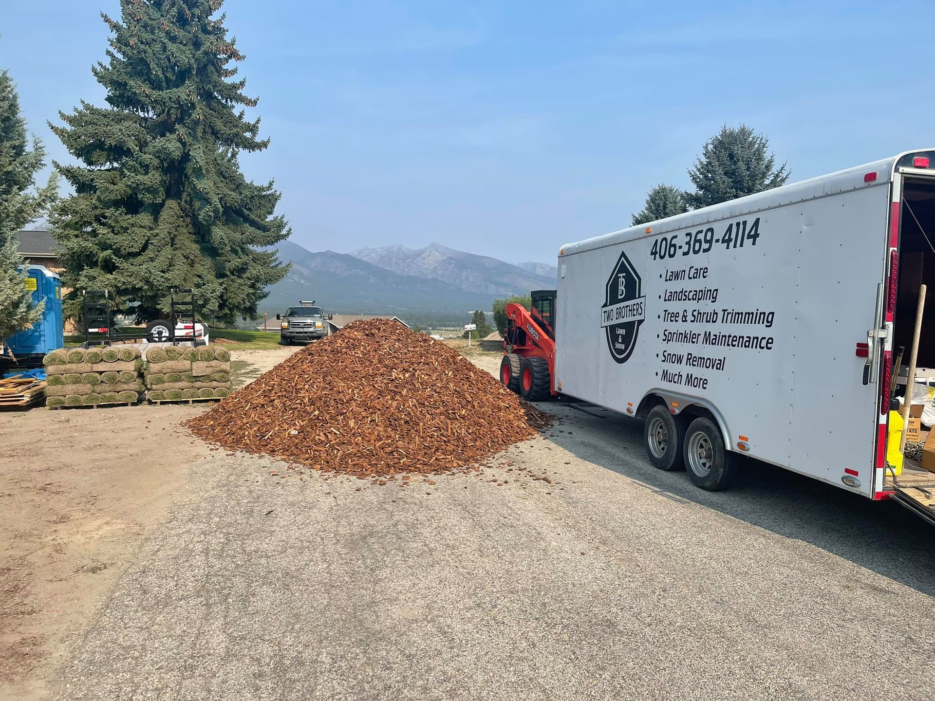 A pile of wood chips next to an open trailer with a company logo, in front of a mountain.