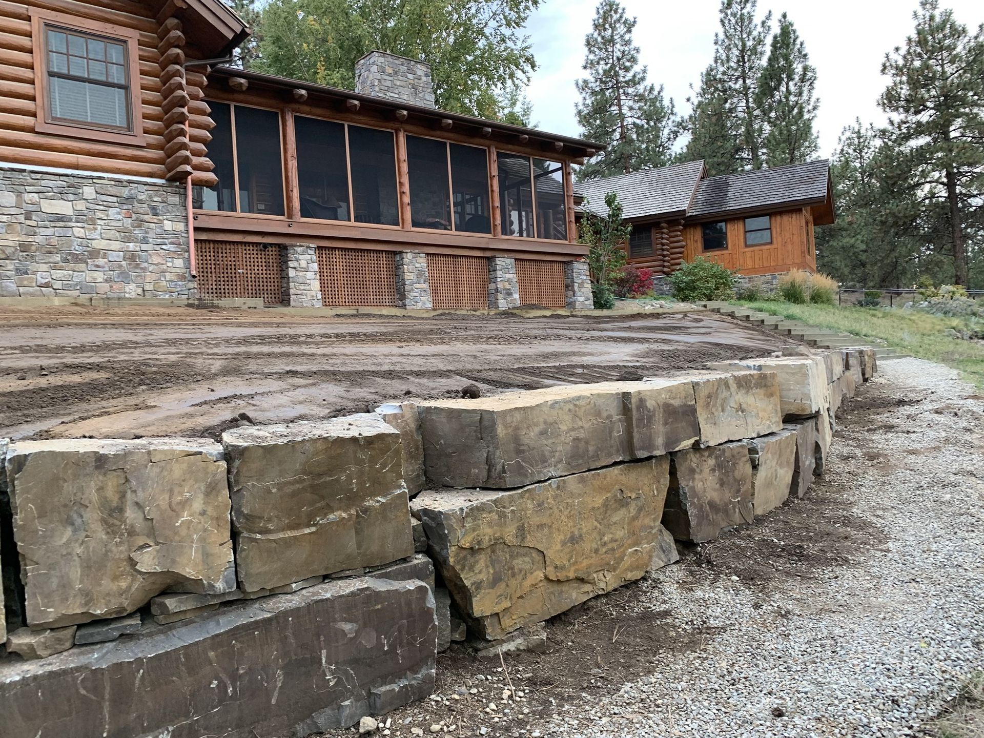 Stone retaining wall in front of a log cabin with a screened porch and small outbuilding in the background.