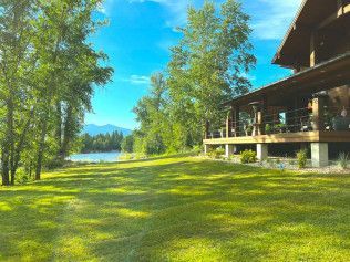 Lush green lawn with trees, lake, and a wooden house under a bright blue sky.