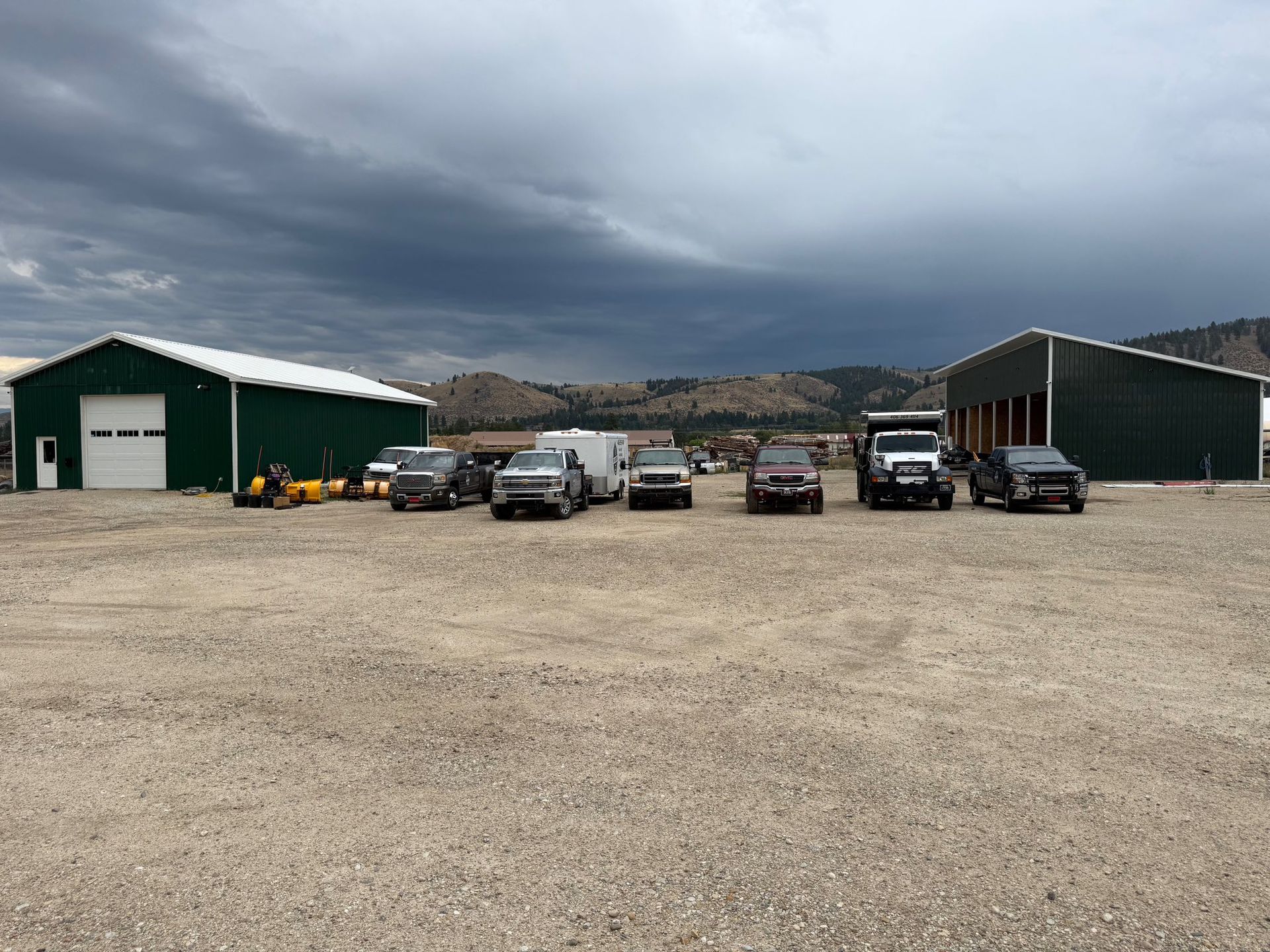 A gravel lot with several vehicles parked in front of two green metal buildings under a cloudy sky.