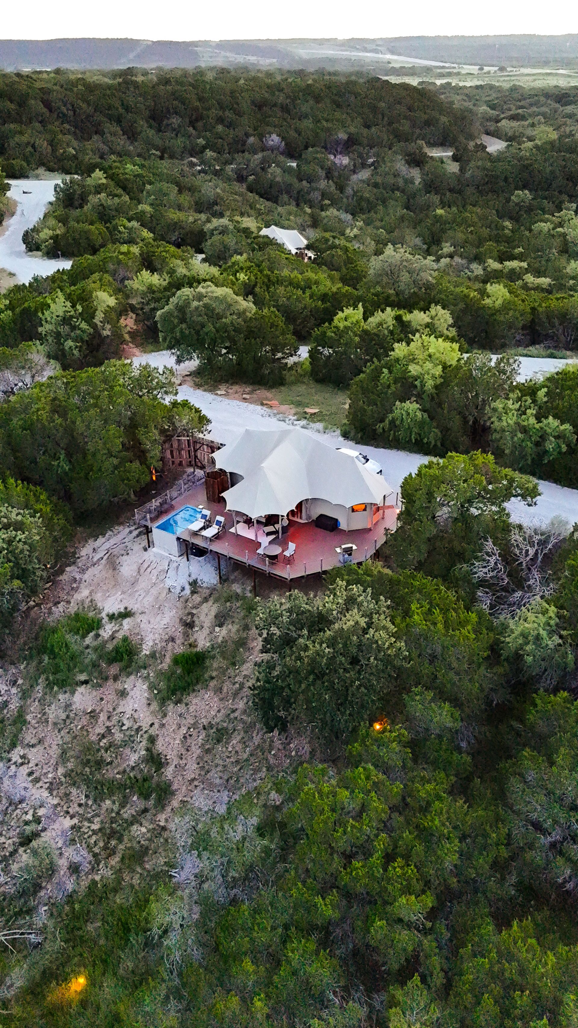 Aerial view of a unique, round house with a wraparound deck surrounded by lush green trees and a winding road.