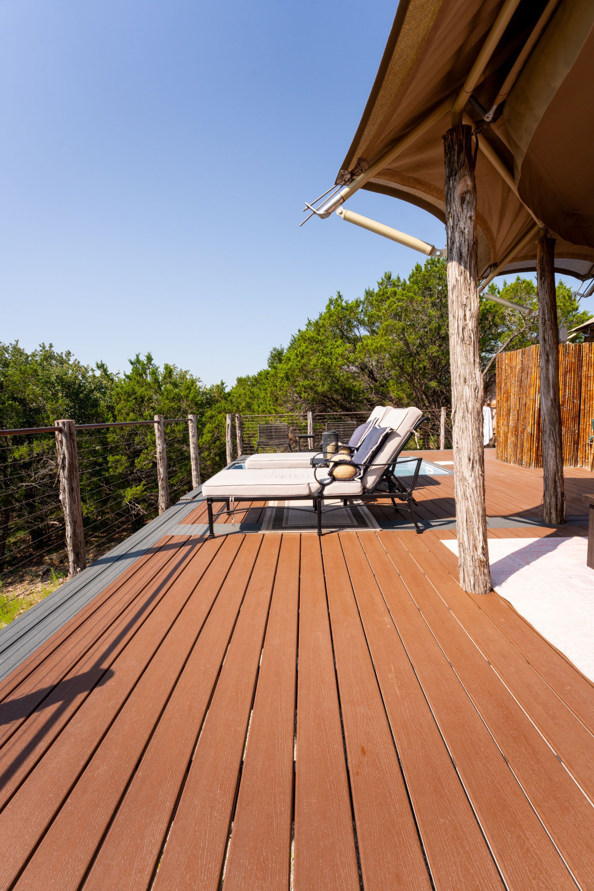 Wooden deck with two lounge chairs under a canvas canopy, overlooking lush green trees against a blue sky.