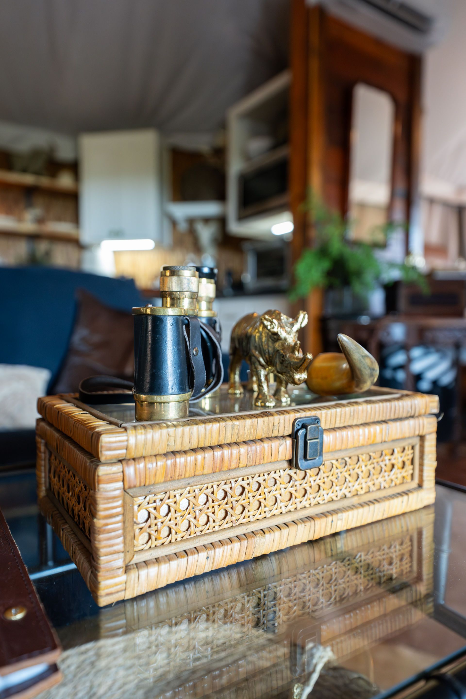 Woven box with brass elephant, binoculars, and other objects on a glass table. Interior, blurred background.