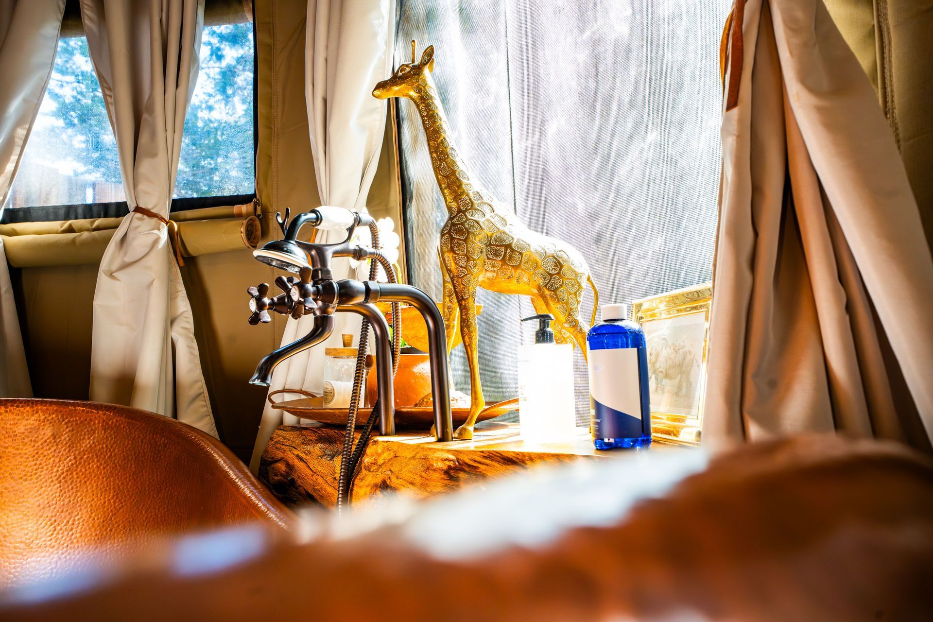 Bathroom shelf with a gold giraffe statue, chrome faucet, and blue bottle in front of a window with curtains.