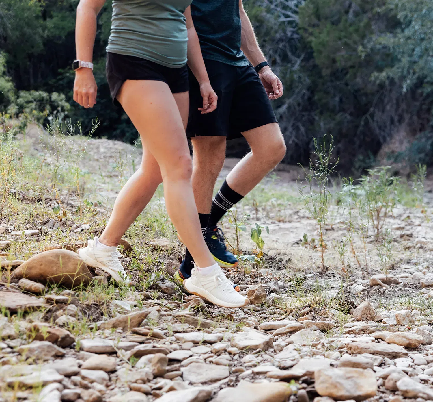 Two people walking outdoors on a rocky path; one wears a green shirt and shorts, the other a black shirt and shorts.