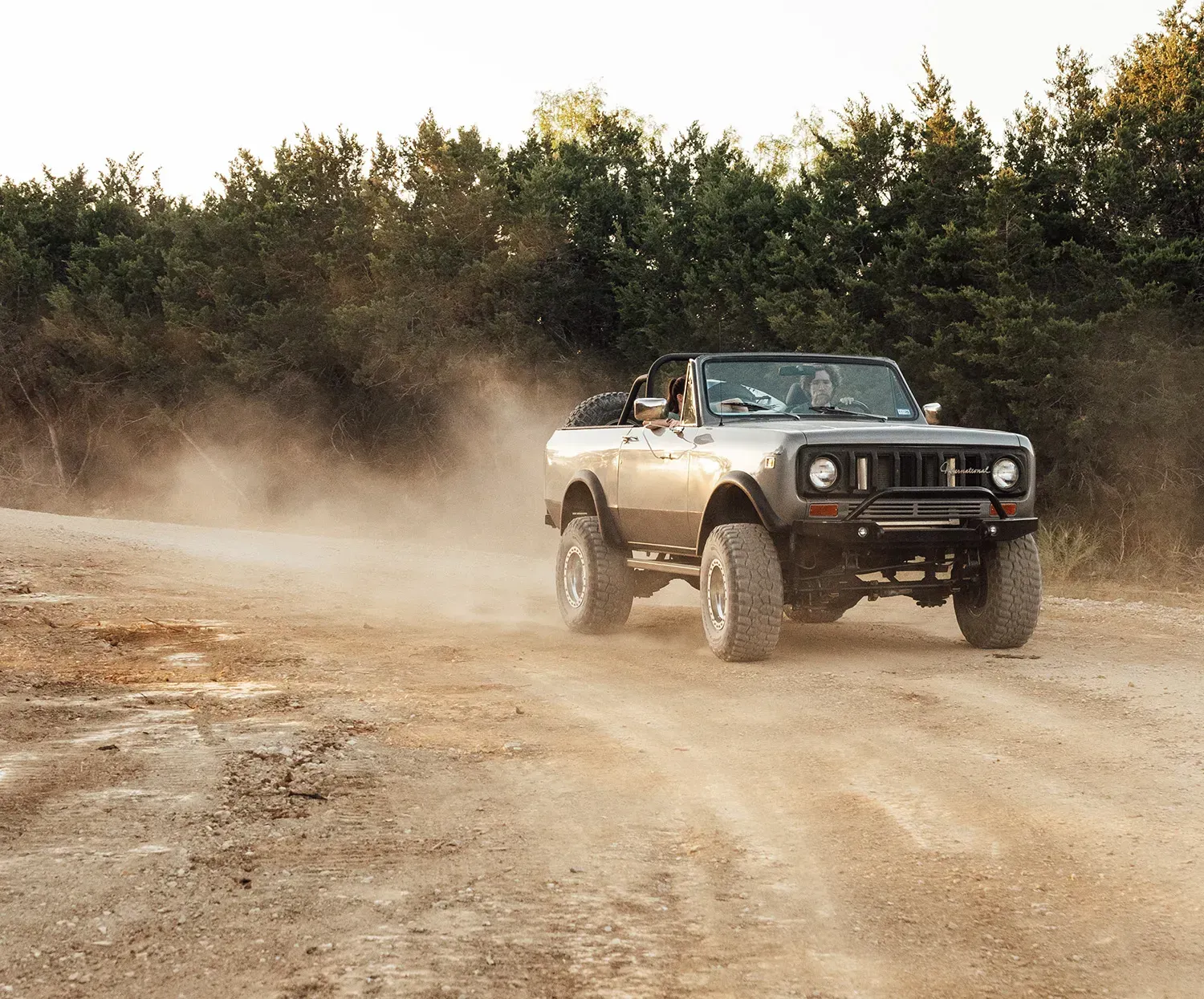 Off-road SUV driving on a dirt road, kicking up dust in front of a tree line.