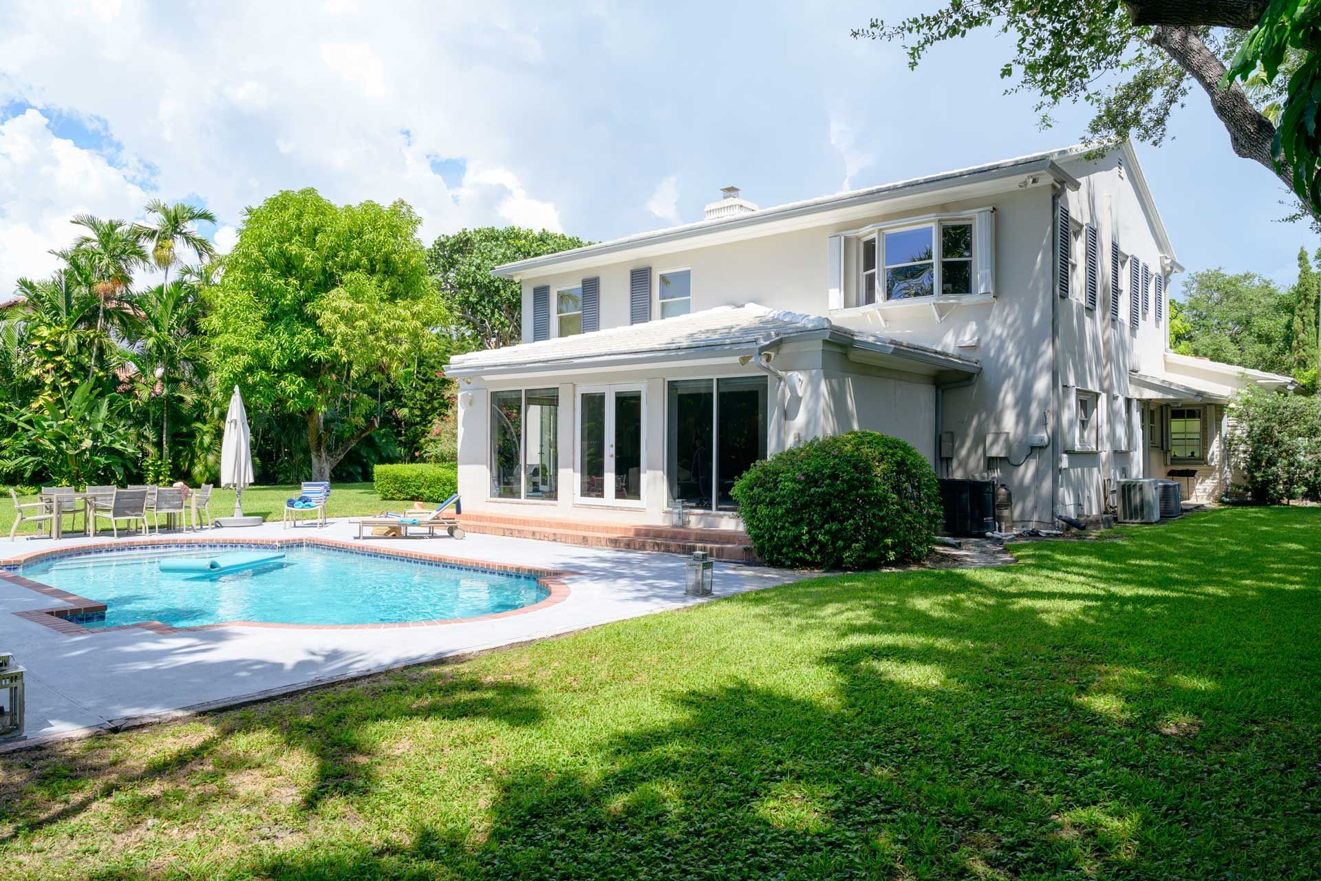 Backyard with swimming pool, green lawn, and two-story white house on a sunny day.