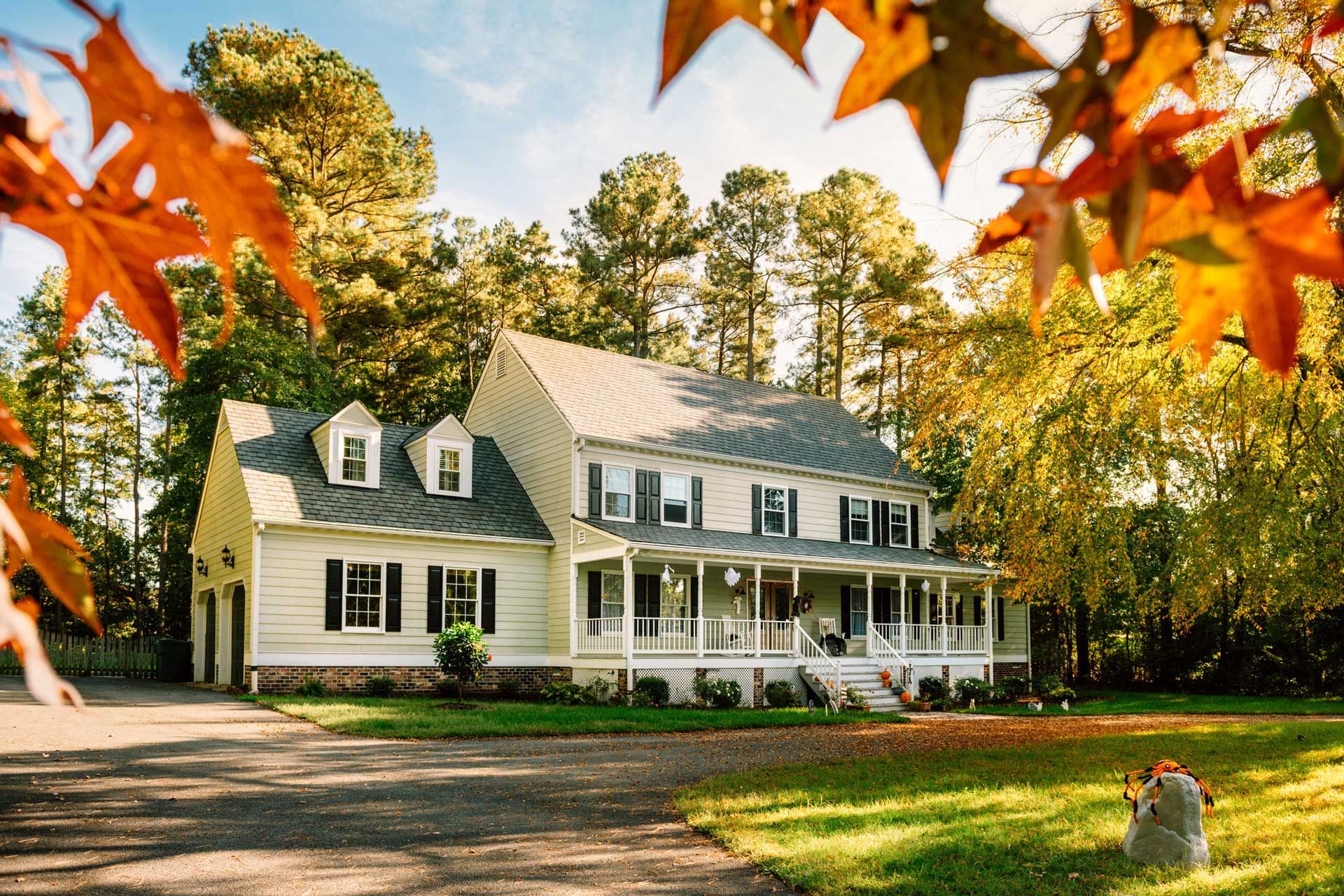 Two-story white house with a porch and dormers, autumn leaves frame the view.
