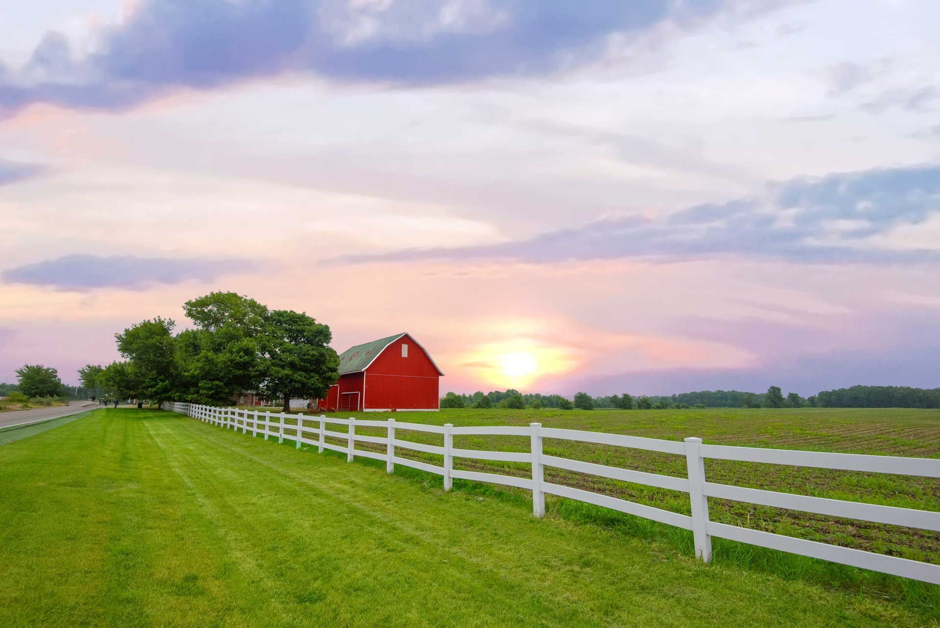 Red barn and white fence in a green field at sunset.