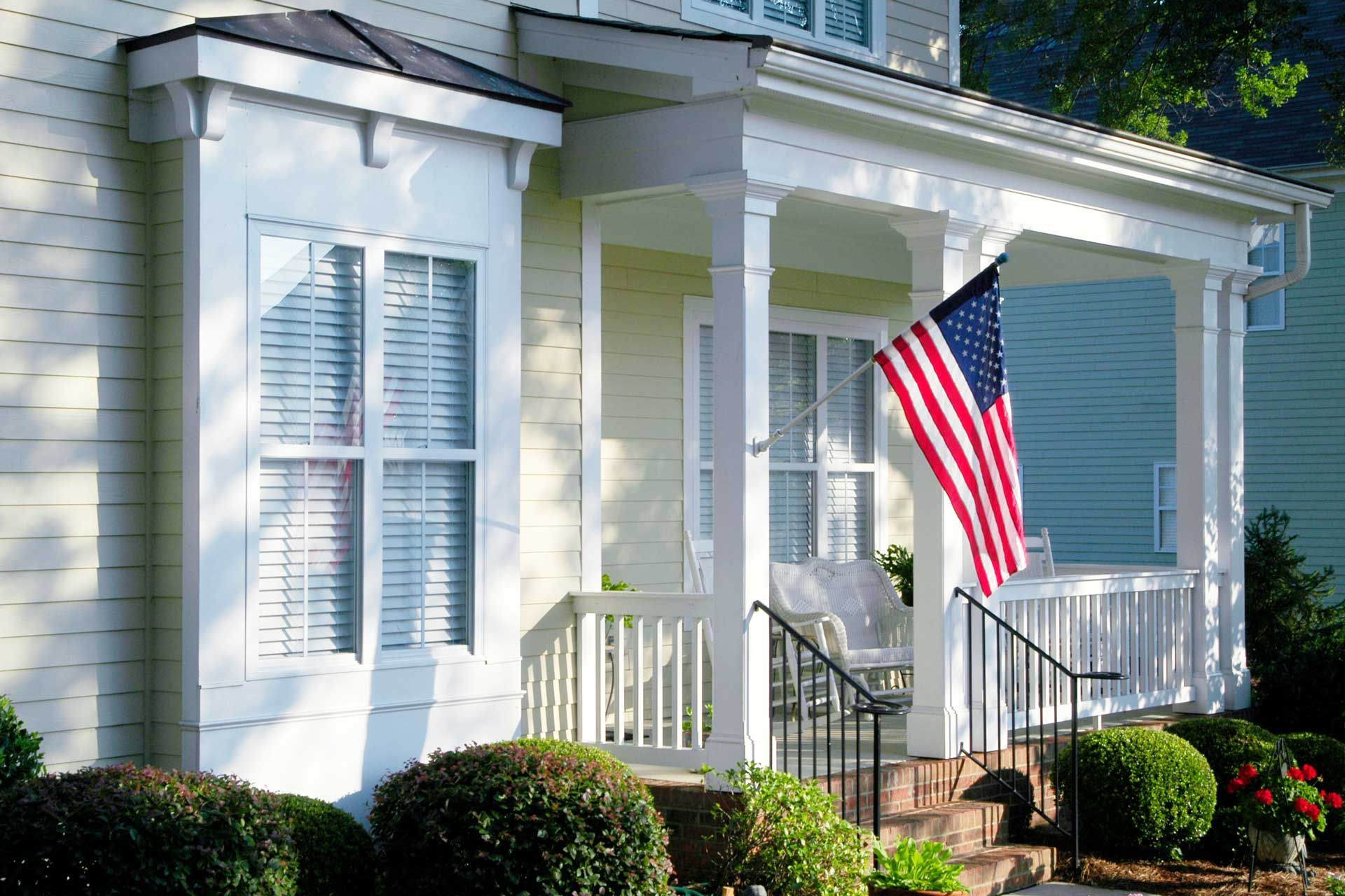 House front with porch, US flag, white trim, bay window, bushes.