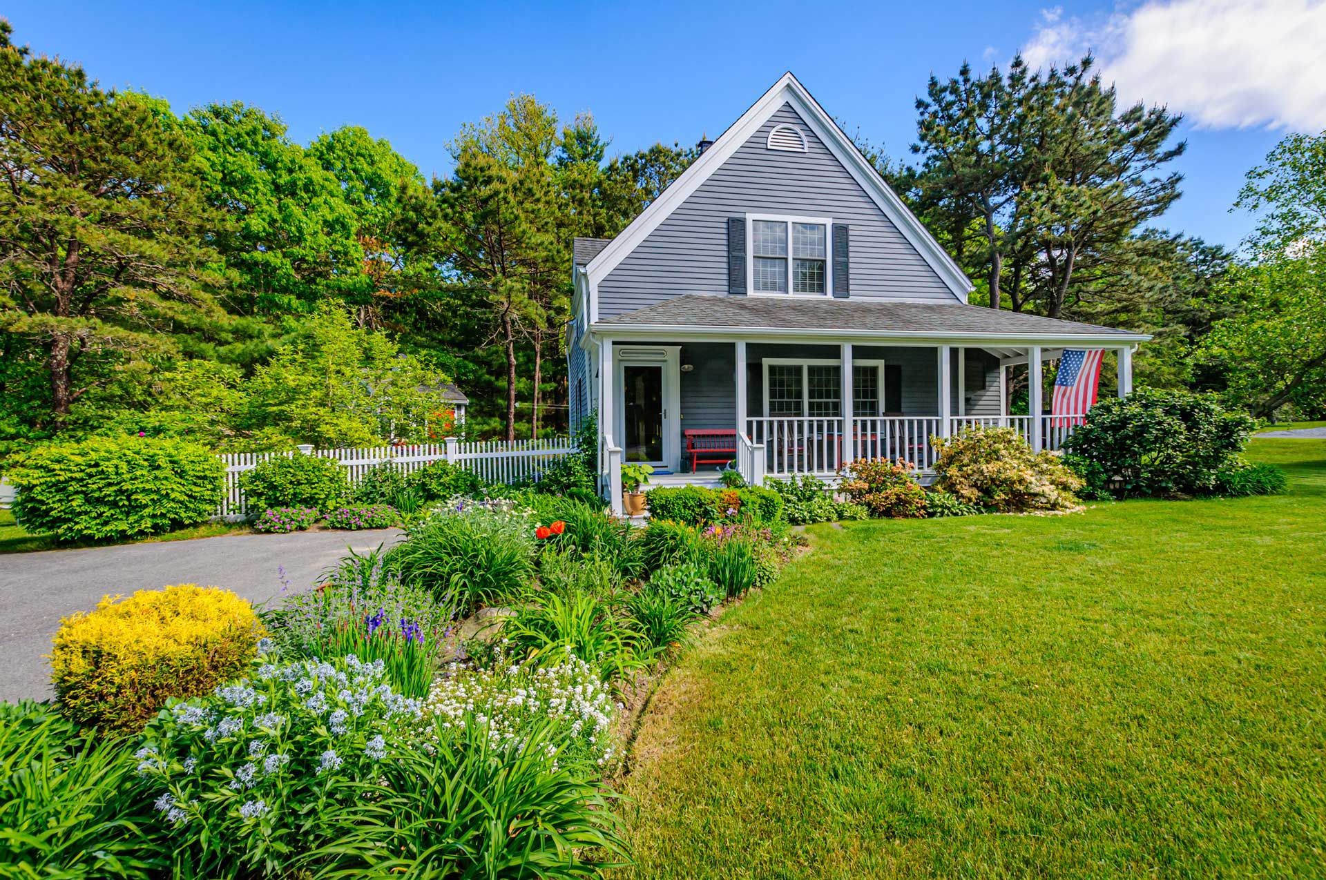 Gray house with white porch and garden with colorful flowers and green lawn.