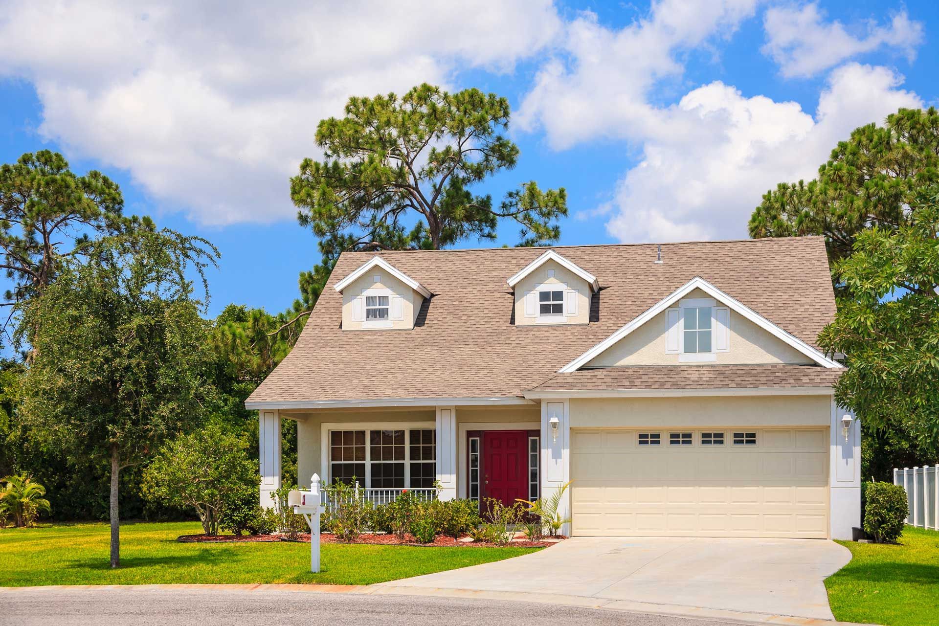 Tan house with red door and white trim, two dormers, attached garage, blue sky, and green trees.