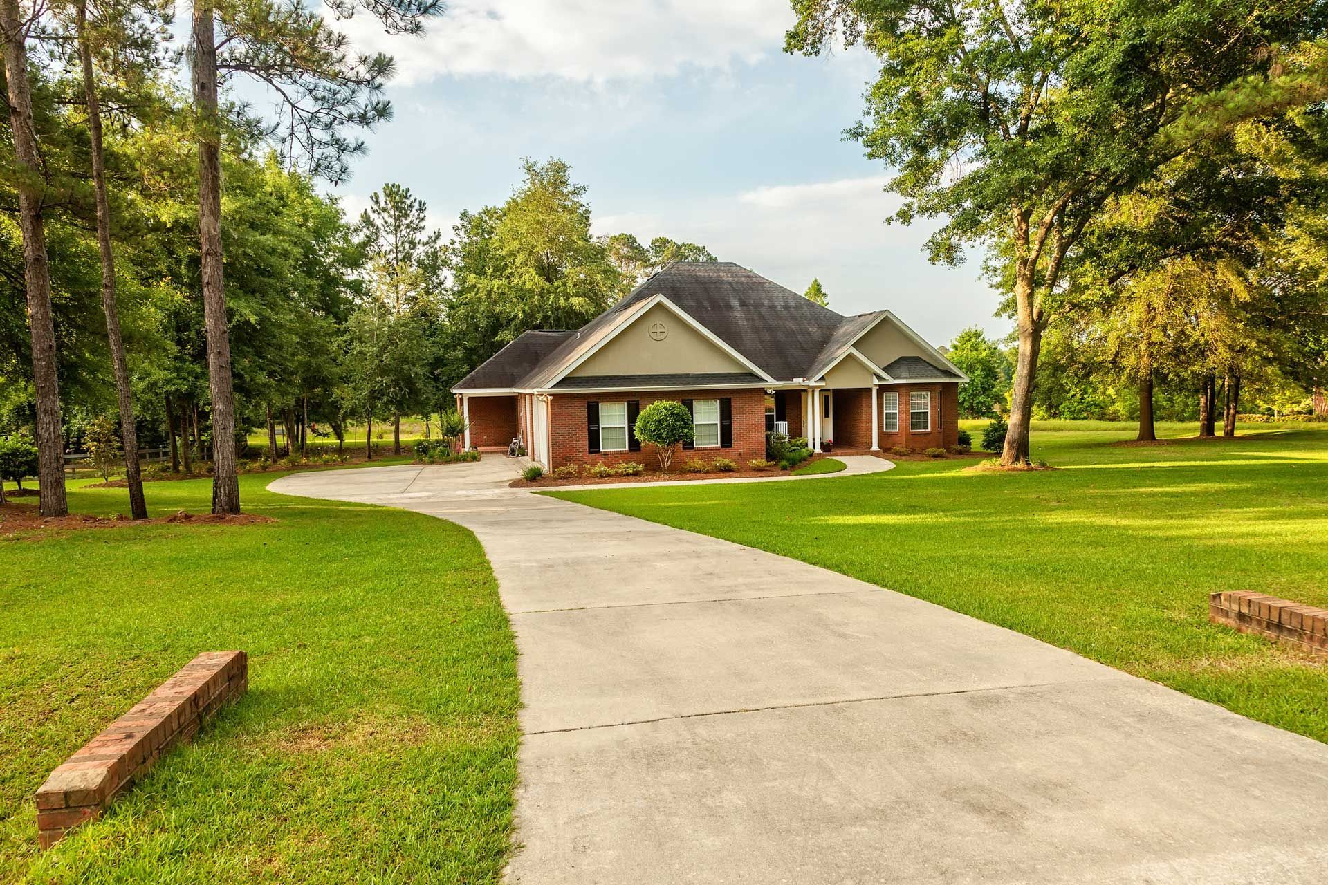 House with long concrete driveway, surrounded by green lawn and trees, under a partly cloudy sky.