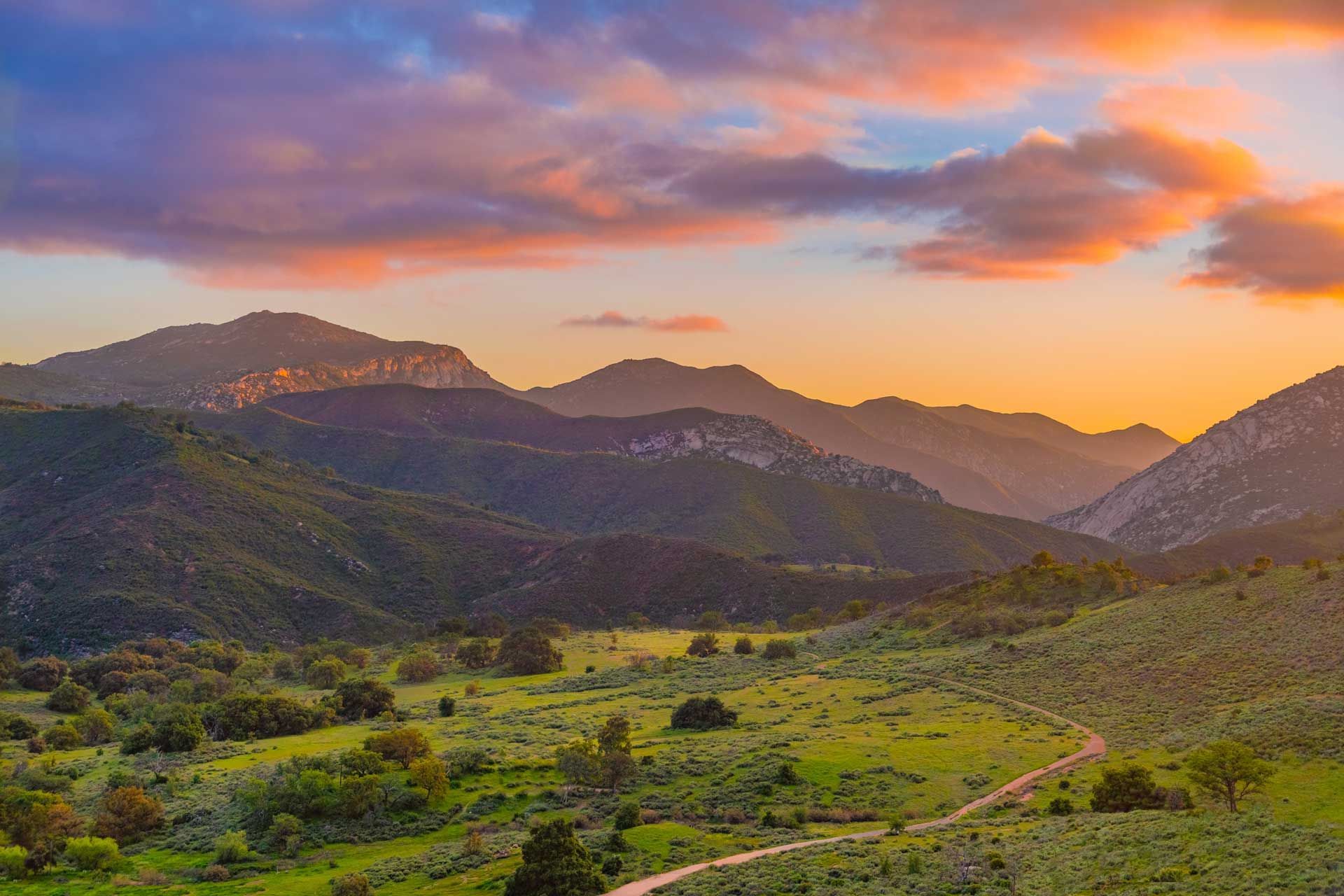Mountains at sunset, with a path winding through a green valley under a sky of orange, pink, and blue hues.
