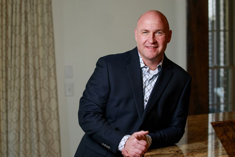 Bald man in navy blazer, leaning on a countertop, smiling. Indoors, natural light.