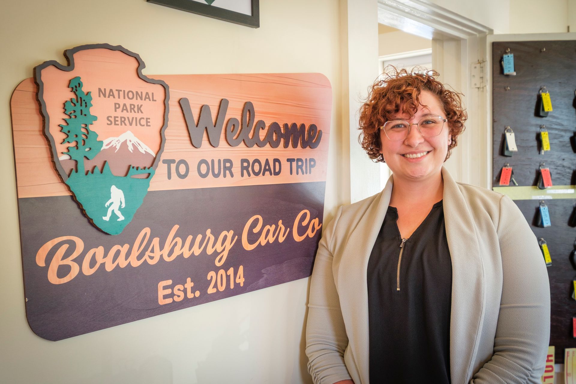 Woman smiling next to a wooden sign for Boalsburg Car Co. with a National Park Service logo. | Boalsburg Car Co.