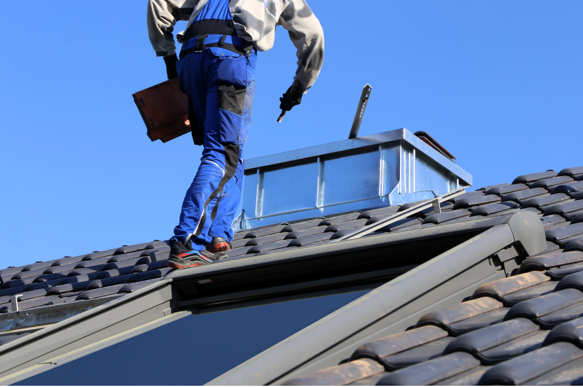 A man in blue overalls is standing on top of a roof.