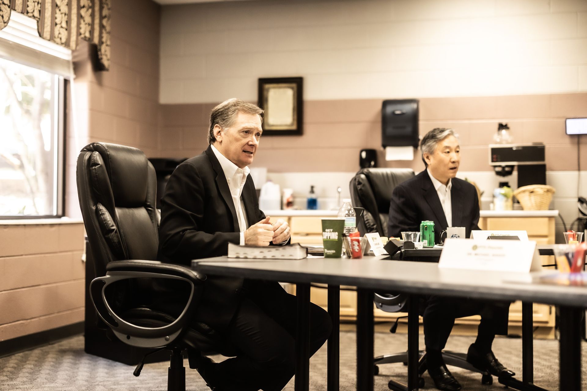 Two men in suits are sitting at a table in a room.