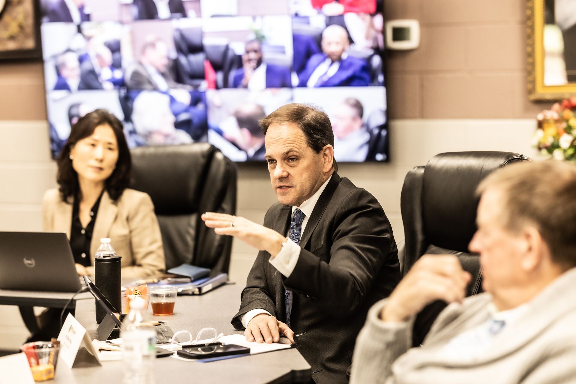 A man in a suit and tie is sitting at a table with other people in a conference room.