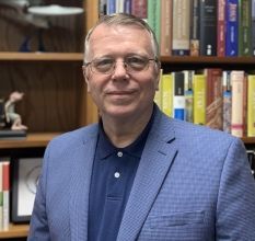 A man in a suit and glasses is standing in front of a bookshelf.