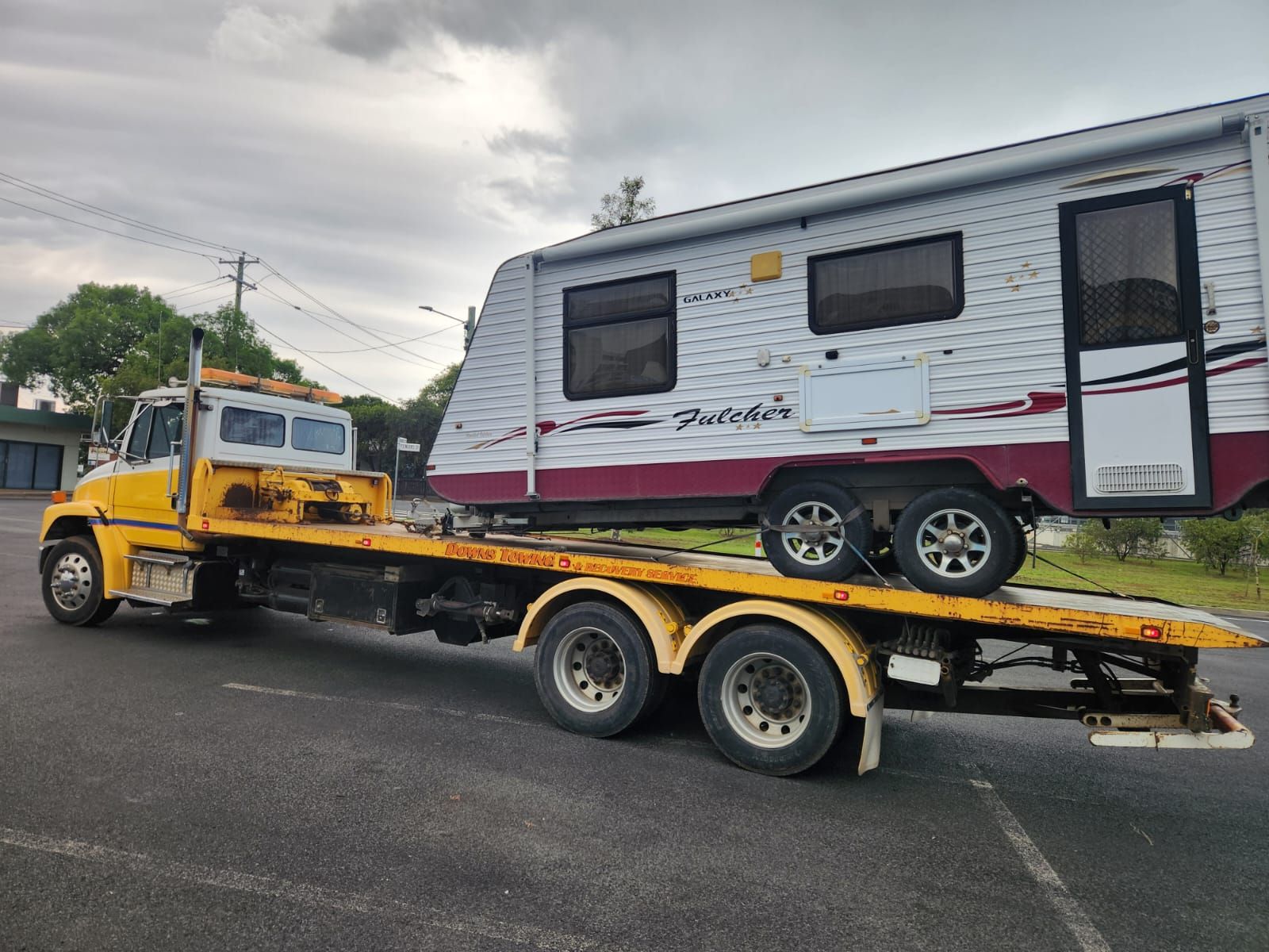 Yellow tow truck transporting a white and red travel trailer on a paved road. Cloudy sky — Downs Towing & Recovery Service in Chinchilla, QLD