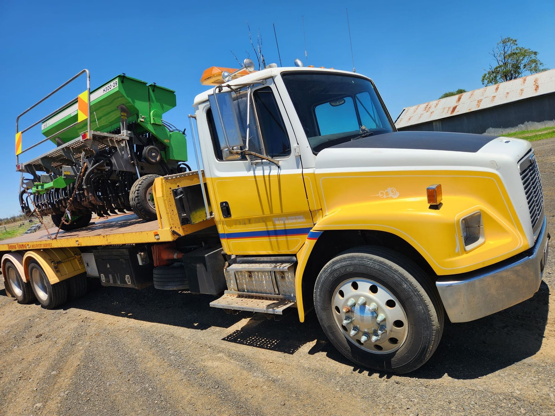 Yellow and white flatbed truck carrying agricultural equipment on a dirt road  — Downs Towing & Recovery Service in Dalby, QLD