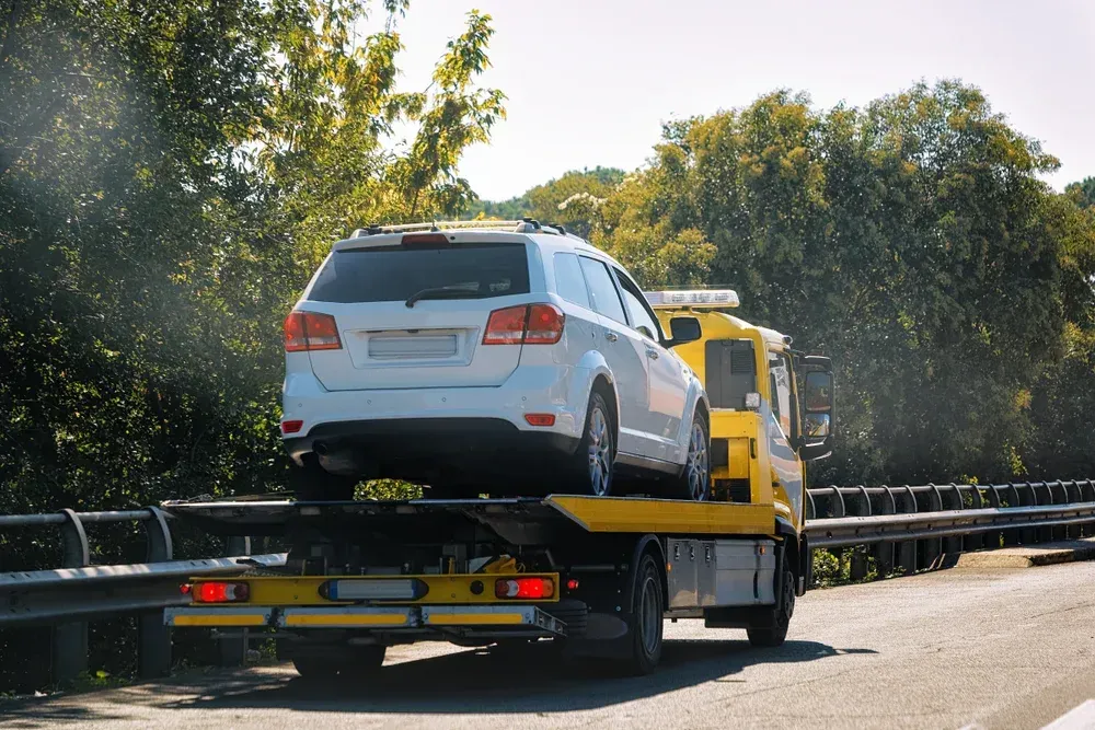 White car being towed on a yellow tow truck, traveling down a road lined with trees  — Downs Towing & Recovery Service in Moonie, QLD
