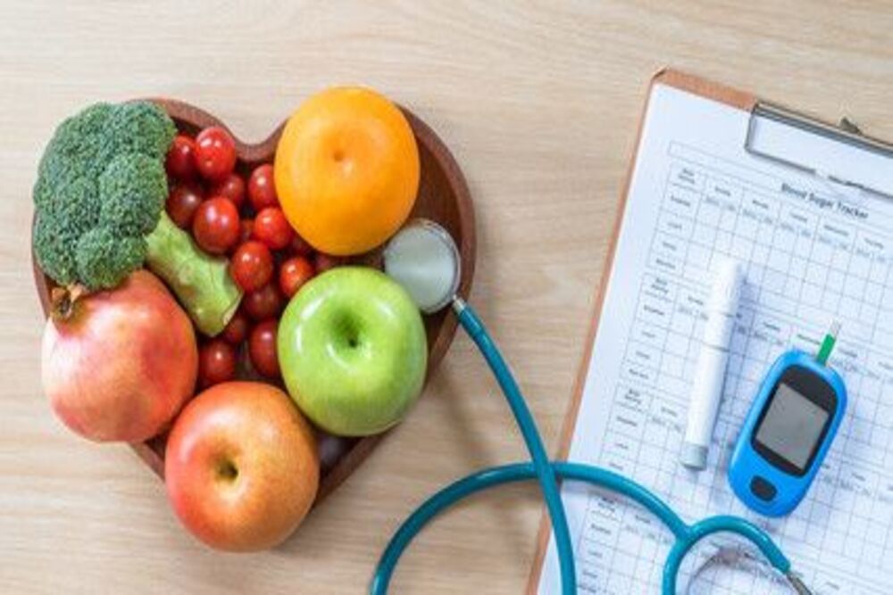 A heart shaped bowl filled with fruits and vegetables next to a stethoscope and a clipboard.