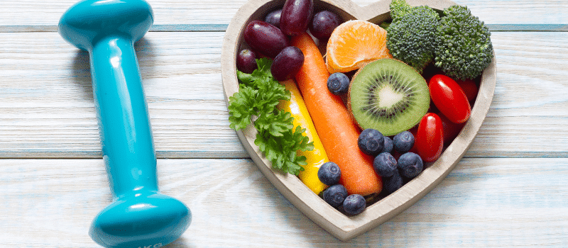 A heart shaped bowl filled with fruits and vegetables next to a blue dumbbell.