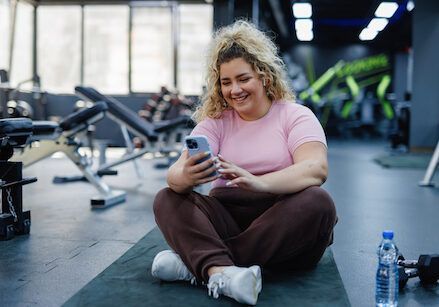 A woman is sitting on a yoga mat in a gym looking at her phone.