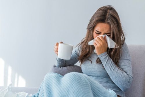 A woman is sitting on a couch blowing her nose while holding a cup of coffee.