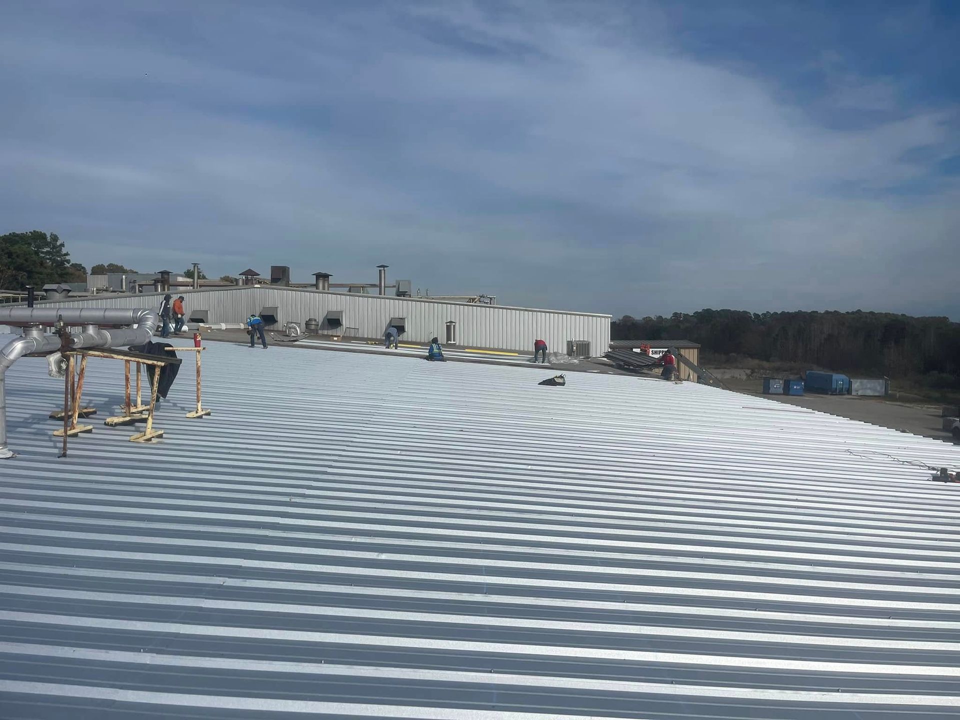 Workers on a large, corrugated metal roof, with a cloudy sky overhead.