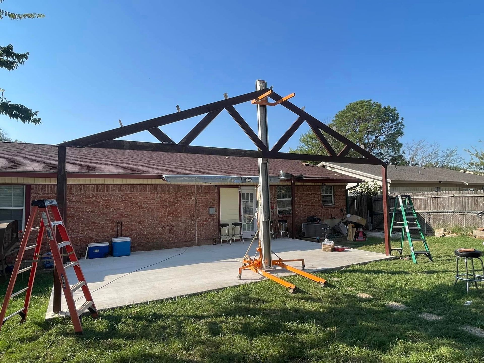 A partially constructed brown metal patio cover supported by a lift on a concrete patio, with a brick house and yard in the background.