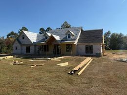 House under construction, with exposed framing and roofing materials, set in a grassy yard under a blue sky.