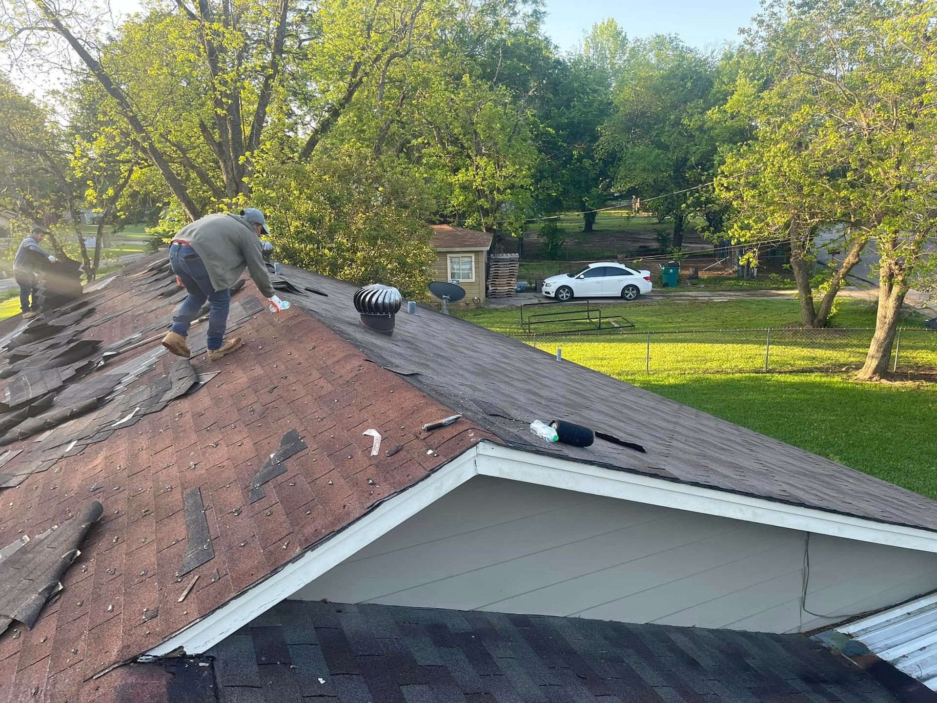 Man repairs roof, shingles are missing. Car and trees in background.