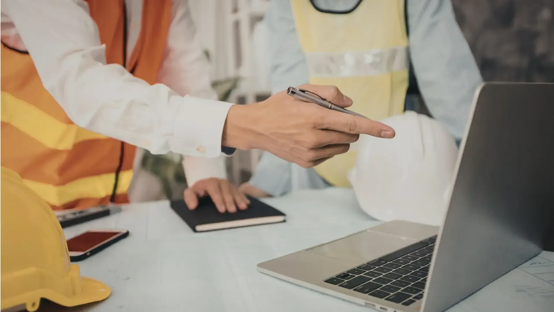 Engineers in safety vests pointing at a laptop screen; helmets, notepad, and phone on table.