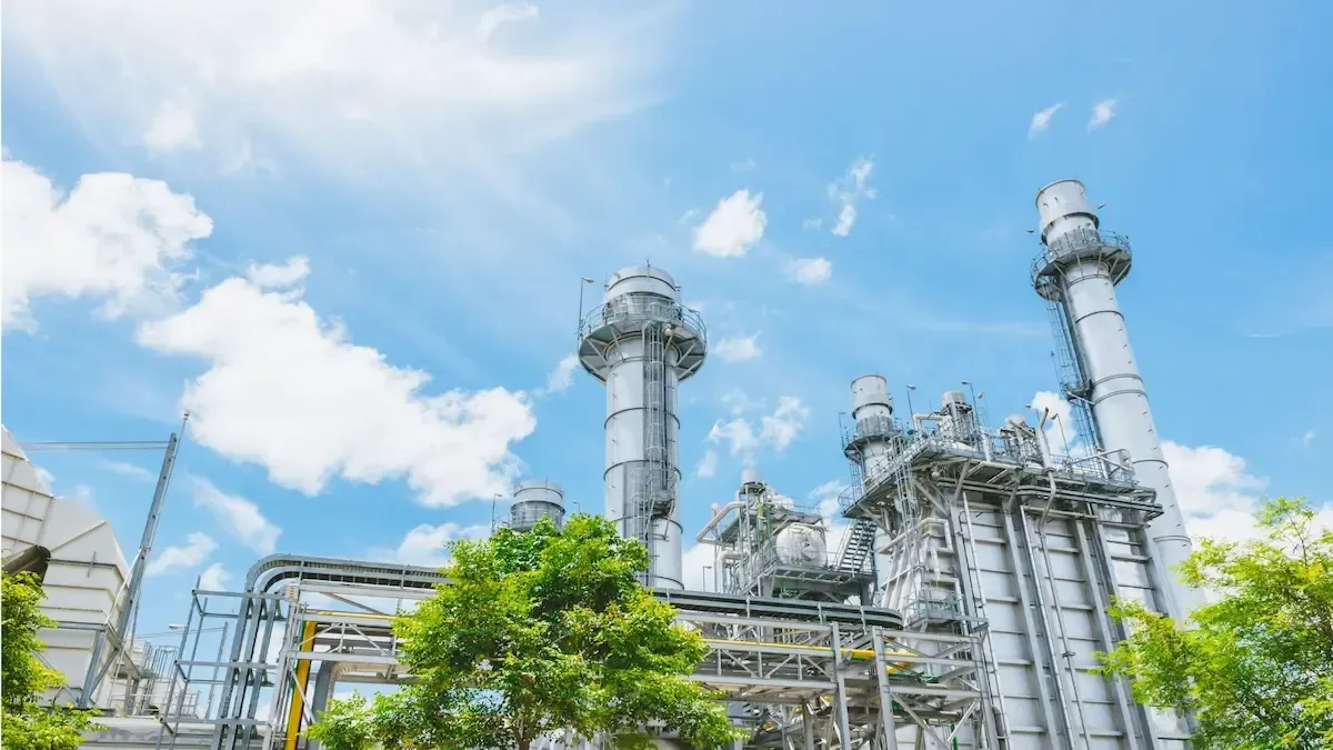 Industrial plant with smokestacks against a bright blue sky, some trees in the foreground.
