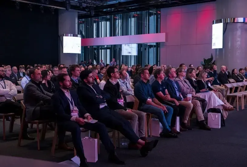 Audience seated in a conference hall, listening to a presentation. People are dressed in business casual attire, with a focus on the speaker.