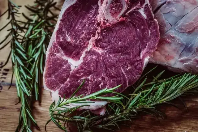 a piece of raw beef is sitting on a wooden cutting board next to rosemary .
