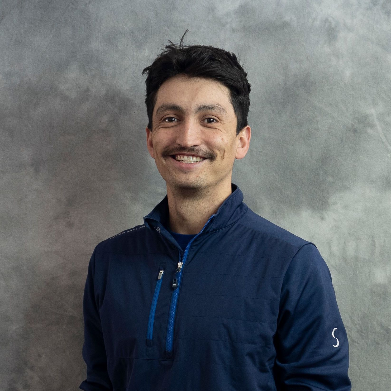 A man with a mustache is wearing a blue jacket and smiling.