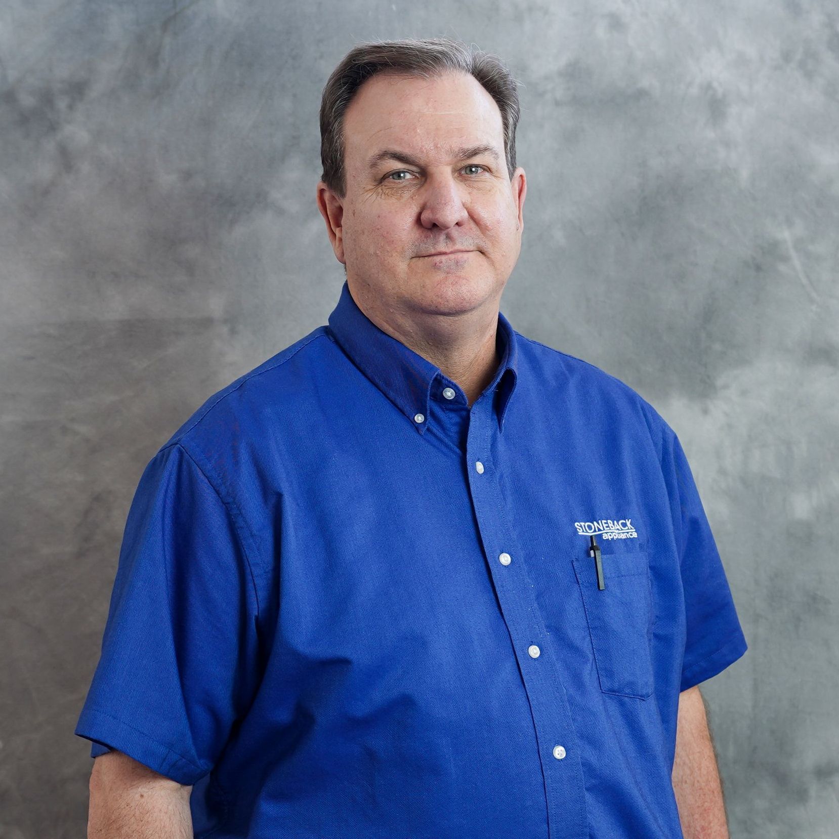 A man in a blue shirt is standing in front of a grey wall.