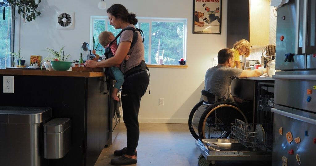 A woman is standing in a kitchen with a baby and a man in a wheelchair.