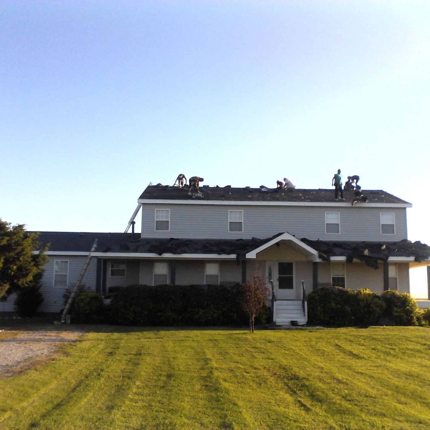 Roofers working on a two-story gray house under a clear blue sky. Green lawn in foreground.
