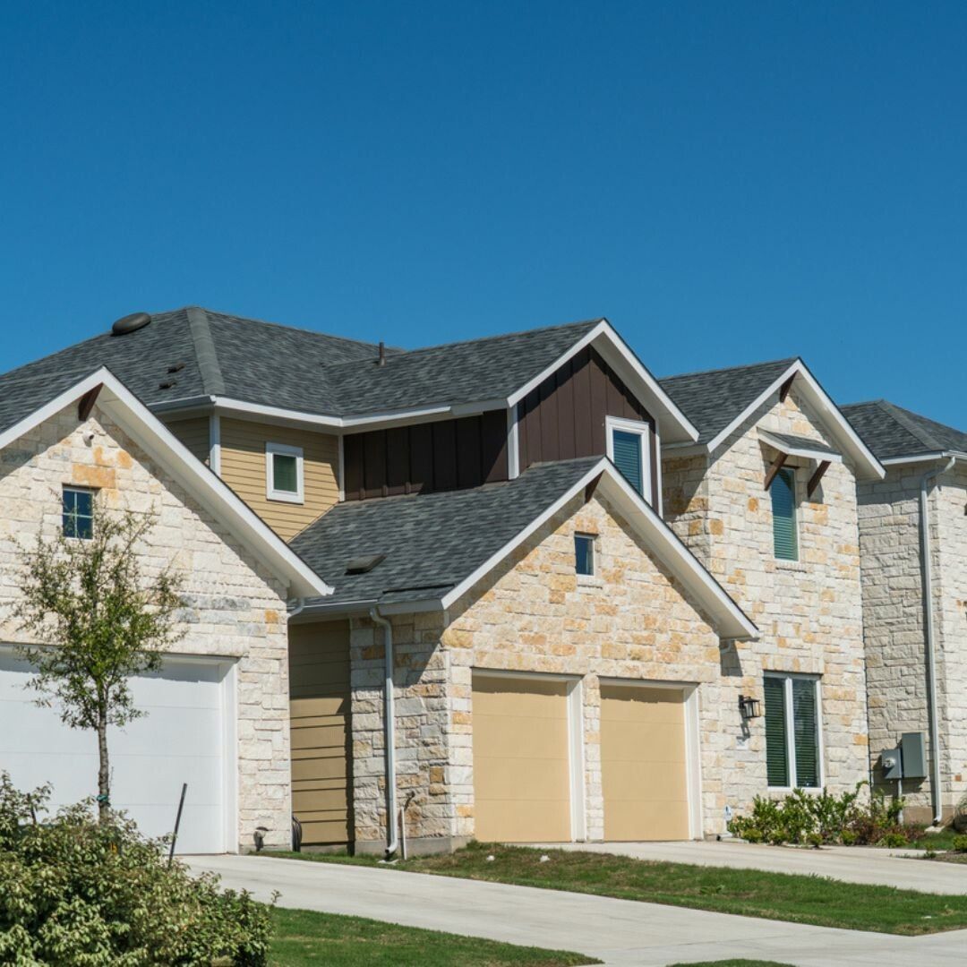 Stone-faced two-story house with multiple gabled roofs, beige garage doors, and a blue sky backdrop.