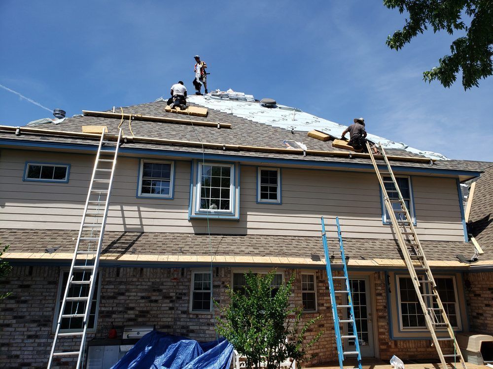 Roofers replacing a shingled roof on a two-story house. Blue sky, ladders, and tarp visible.